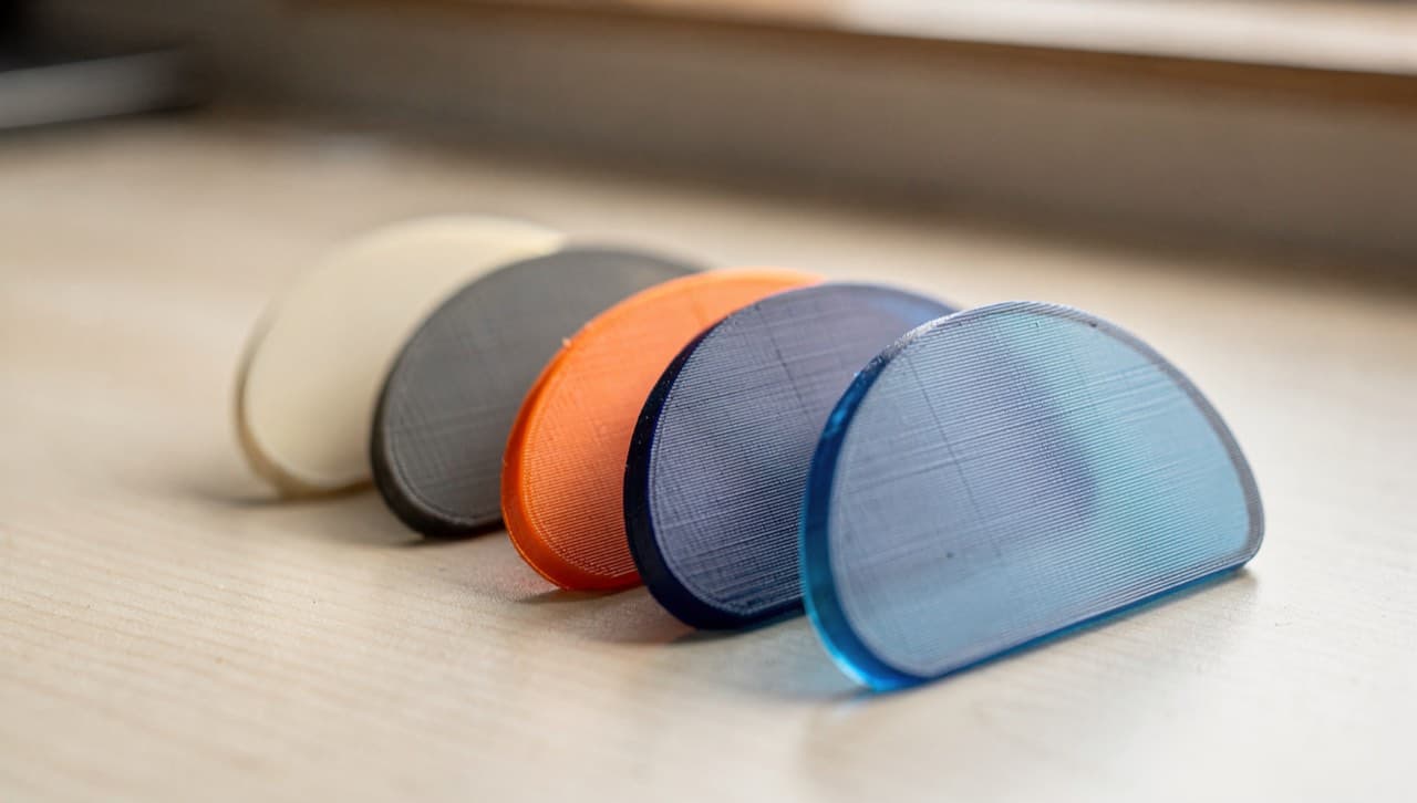 A fanned row of 3D-print colour swatch chips in matte cream, charcoal, filament orange, deep blue, and a translucent blue, laid on a light bench in soft daylight showing how layer lines catch the light differently per colour.