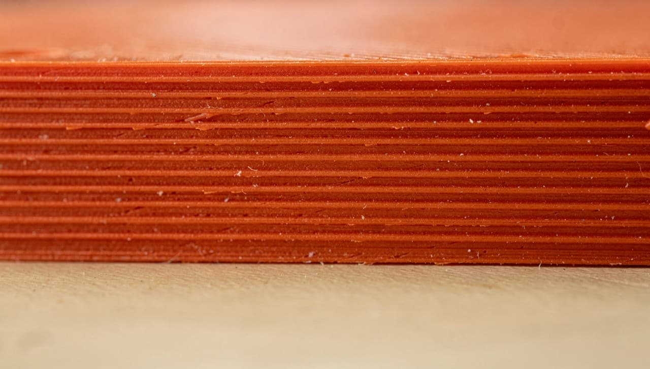 Macro photograph of horizontal layer lines on a matte filament-orange 3D-printed plastic surface, side-lit to show the texture of each 0.2 mm layer.