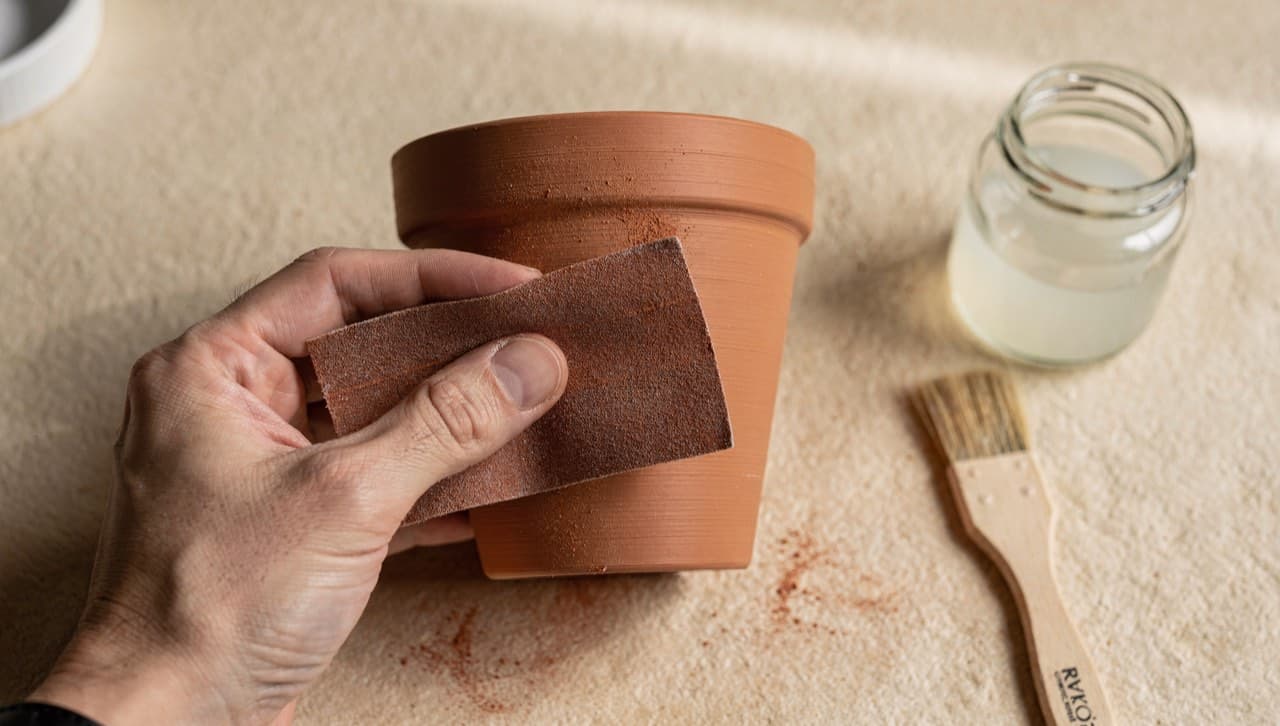 Top-down close-up of a hand mid-finishing a 3D-printed terracotta plant pot with 400-grit sandpaper, with a jar of matte sealer and a brush resting on the workbench beside it.